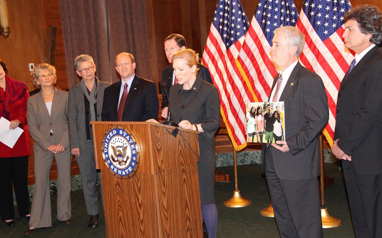 A woman speaks at a United States Senate podium as officials stand behind her with American flags