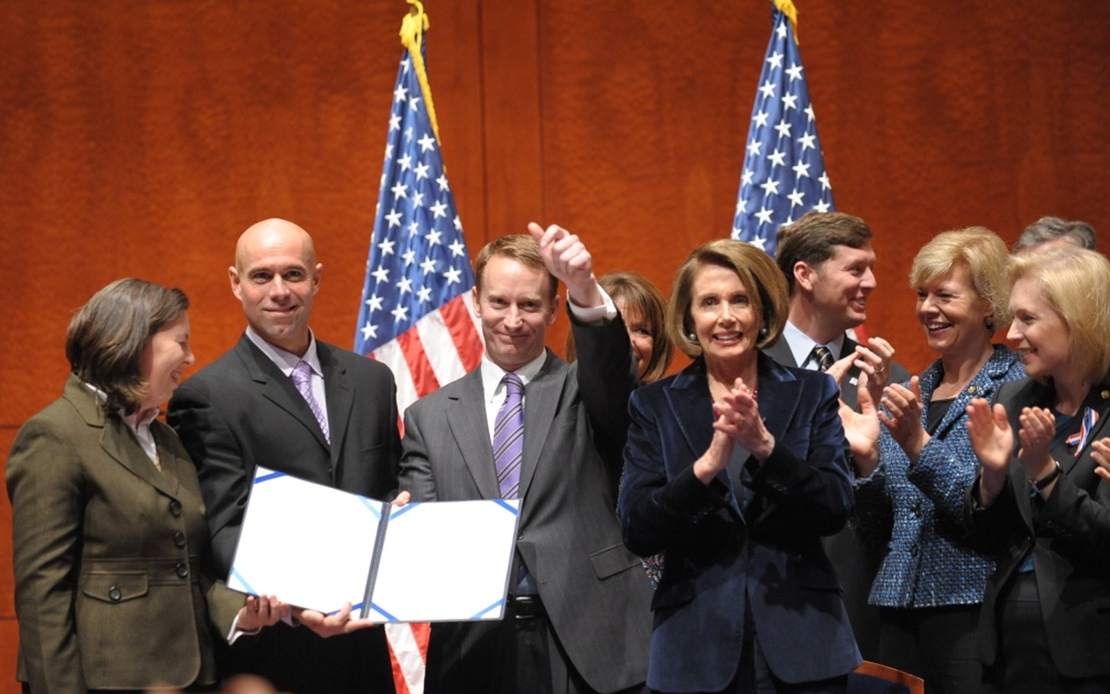 Lawmakers applaud beside a signed document, with American flags in the background
