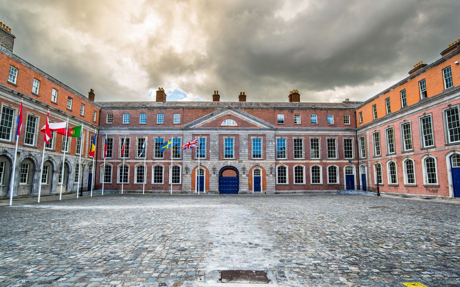 Cobblestone courtyard with flags lined up before a redbrick building under cloudy skies