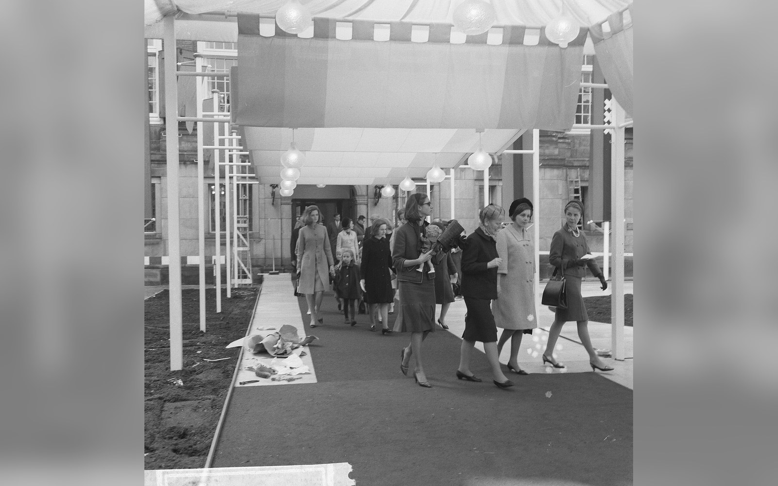 Women and children in coats walk beneath a canopy along a red carpet outside a building