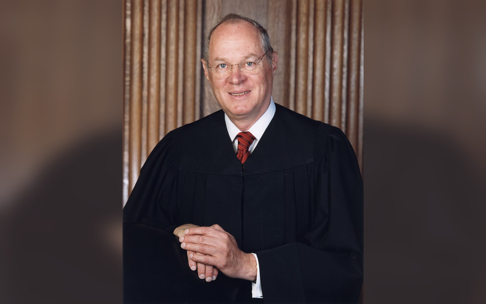 A judge in black robes and red tie poses for an official portrait in front of curtains