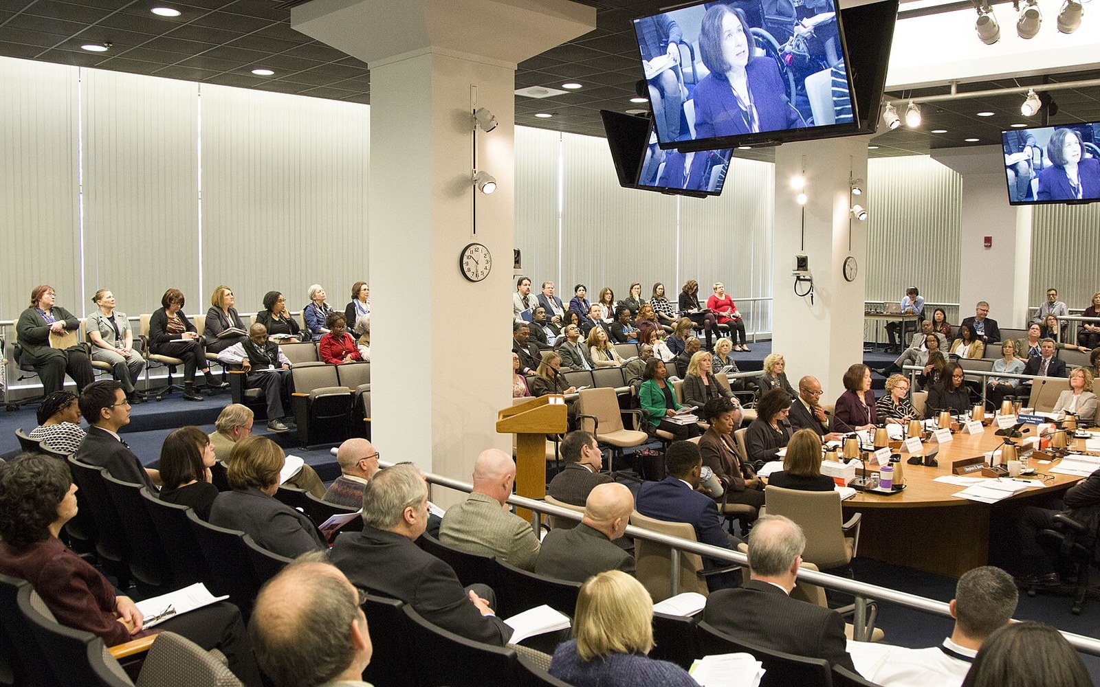 Attendees fill a boardroom for an EEOC briefing, with speakers shown on overhead screens