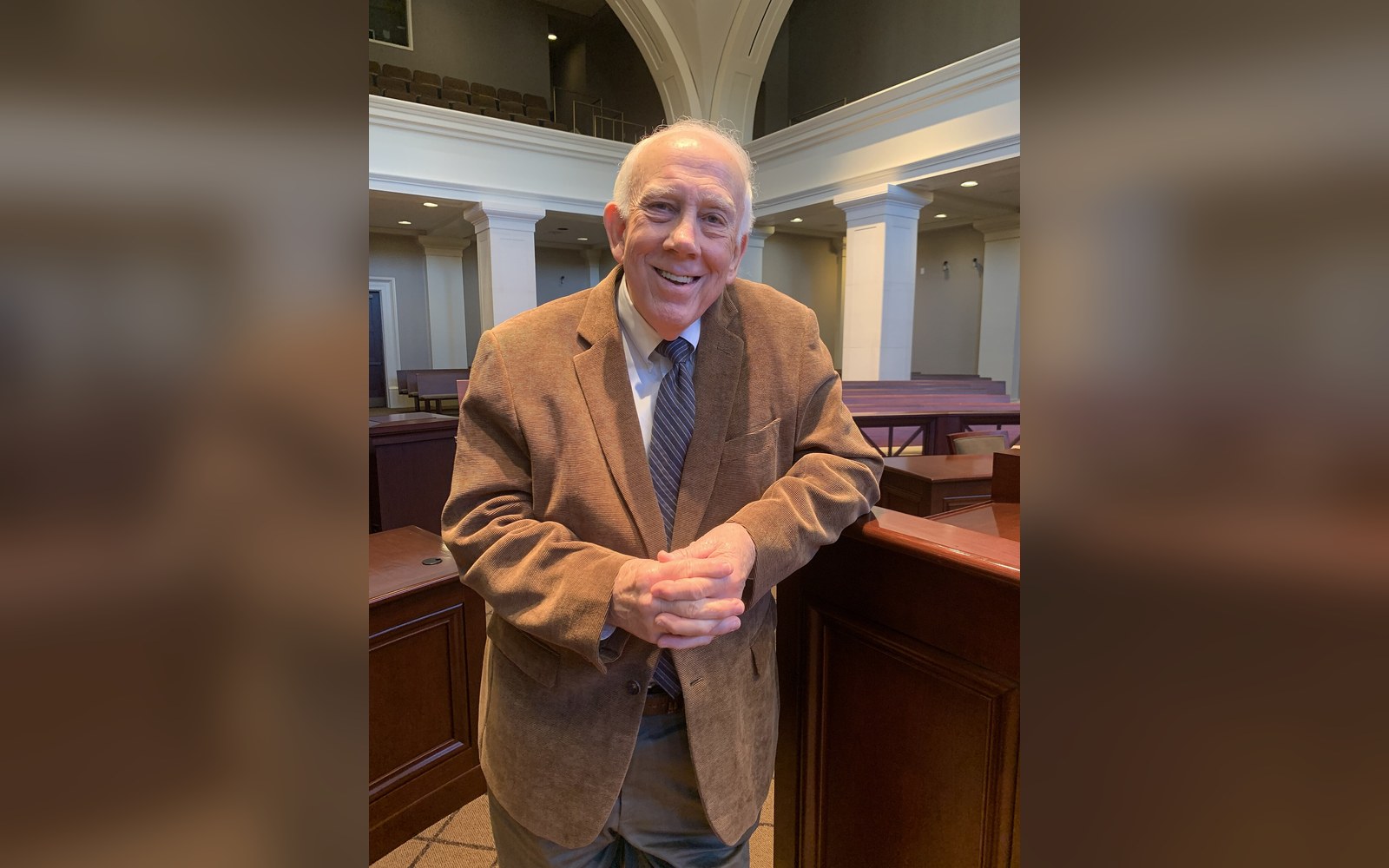 Smiling older man in a tan blazer poses in a wood-paneled courtroom