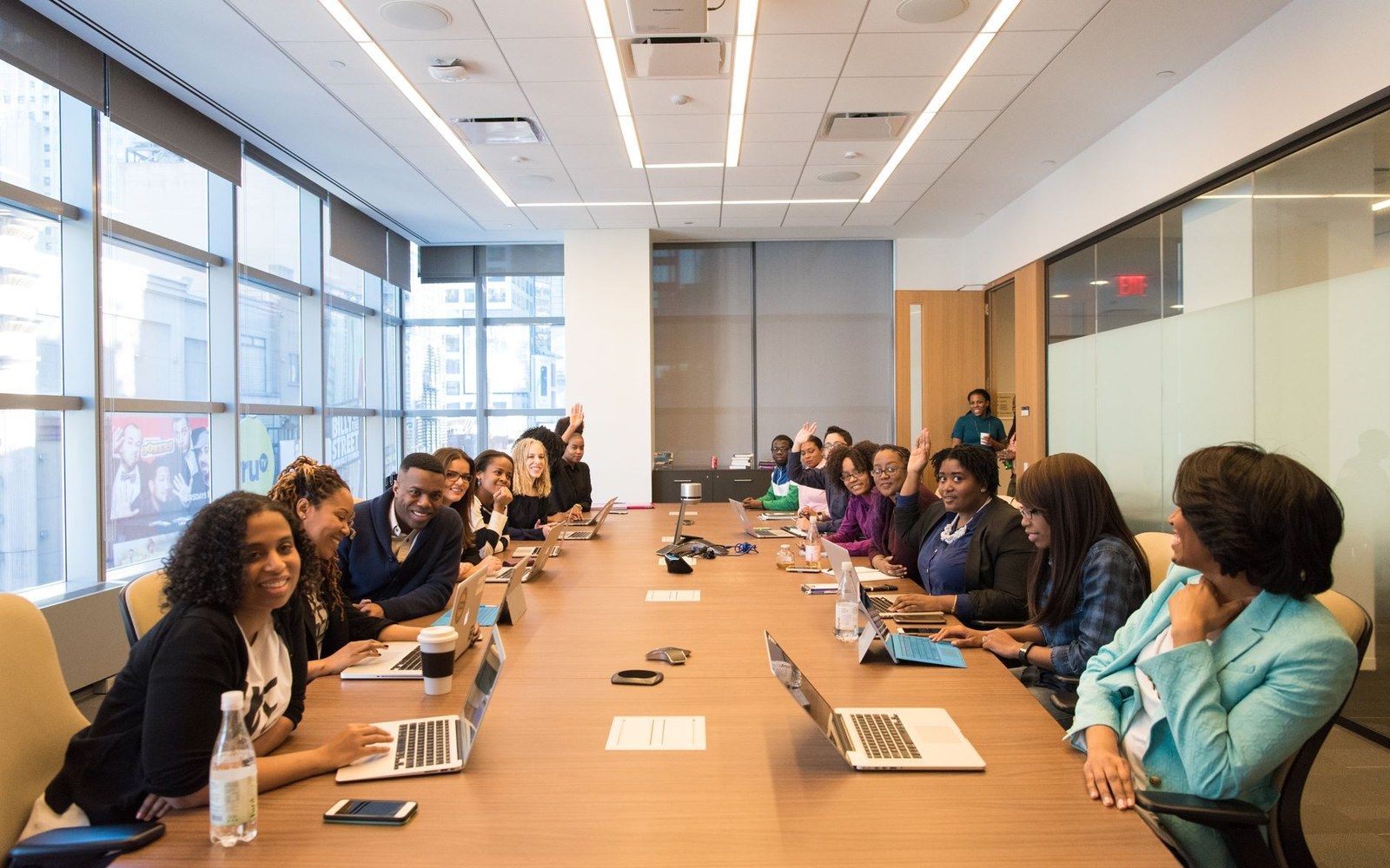 People sit around a long conference table with laptops, some raising their hands during a meeting